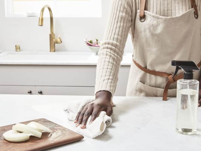 A woman wiping down a counter with rag and clear spray bottle 